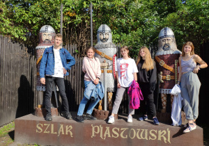Pięcioro uczniów stoi pośród trzech figur rycerzy strzegących Mysiej Wieży. Pod pomnikiem znajduje się napis „Szlak Piastowski”
